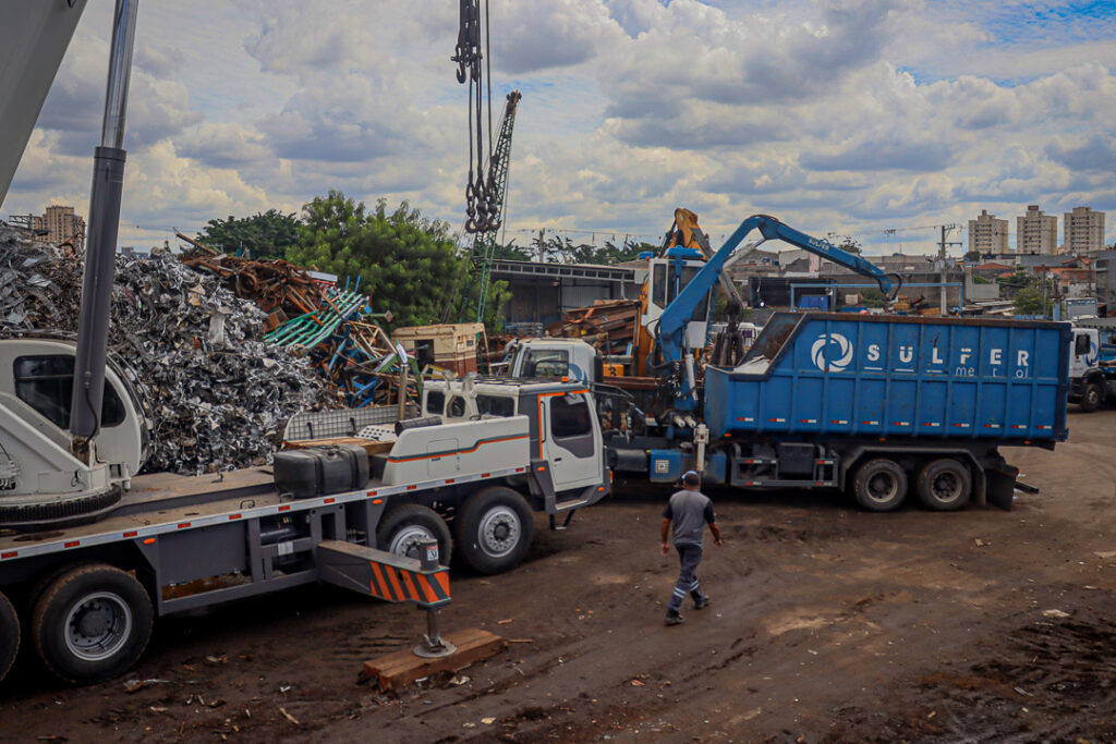 Funcionário da Sulfermetal realizando operação de separação e armazenamento de sucata de estamparia industrial no pátio da empresa. Ao fundo, caminhão munck manipulador de sucata e guindauto em operação, com pilhas de estamparia destinadas à siderurgia visíveis ao redor. Fotografia autoral por Luyza Santos.