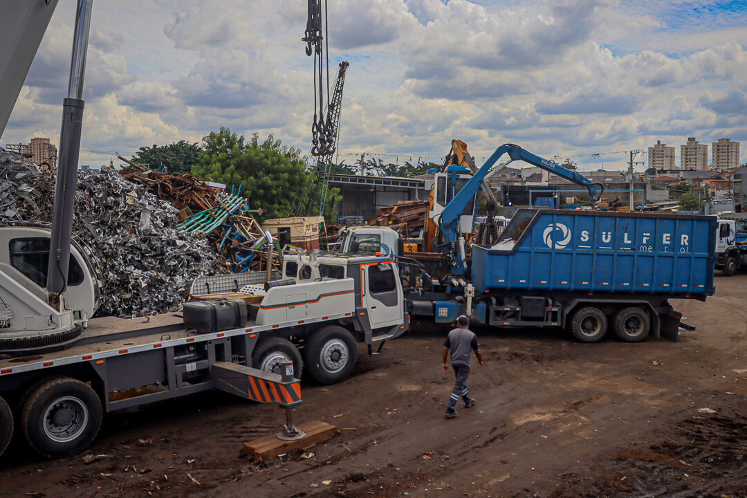Funcionário da Sulfermetal realizando operação de separação e armazenamento de sucata de estamparia industrial no pátio da empresa. Ao fundo, caminhão munck manipulador de sucata e guindauto em operação, com pilhas de estamparia destinadas à siderurgia visíveis ao redor. Fotografia autoral por Luyza Santos.